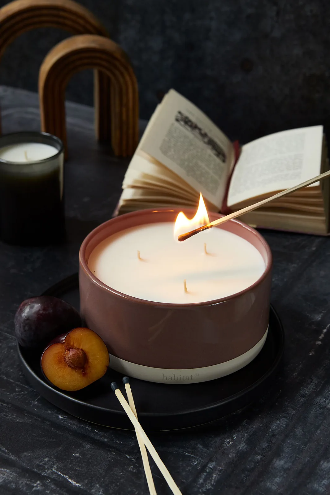 Dark moody product photography of a lit three-wick candle in a terracotta ceramic vessel, styled with an open book, matches, and halved plums on a dark stone surface.