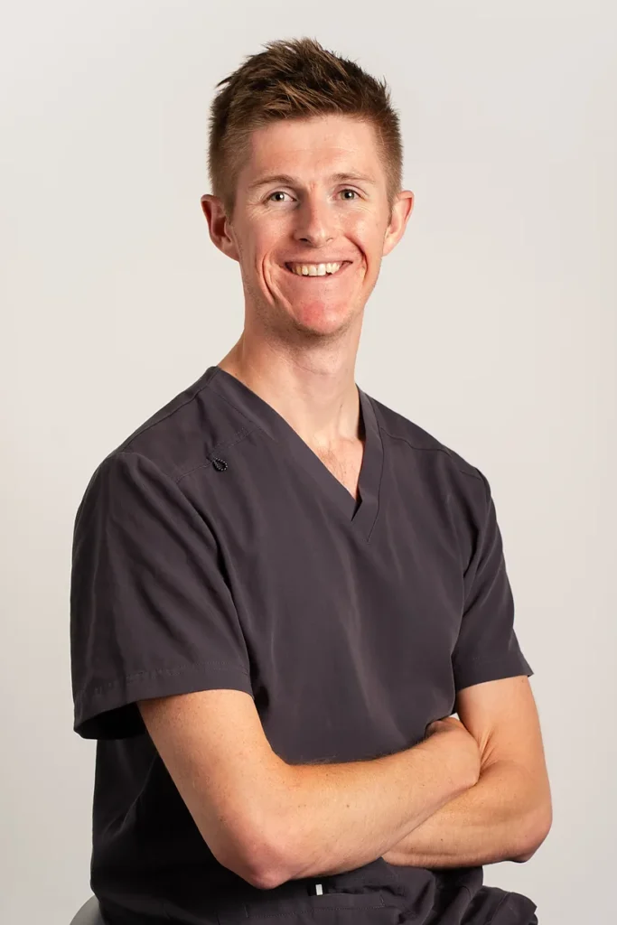 Professional corporate headshot of a smiling man in grey scrubs with arms crossed, against a neutral studio background.