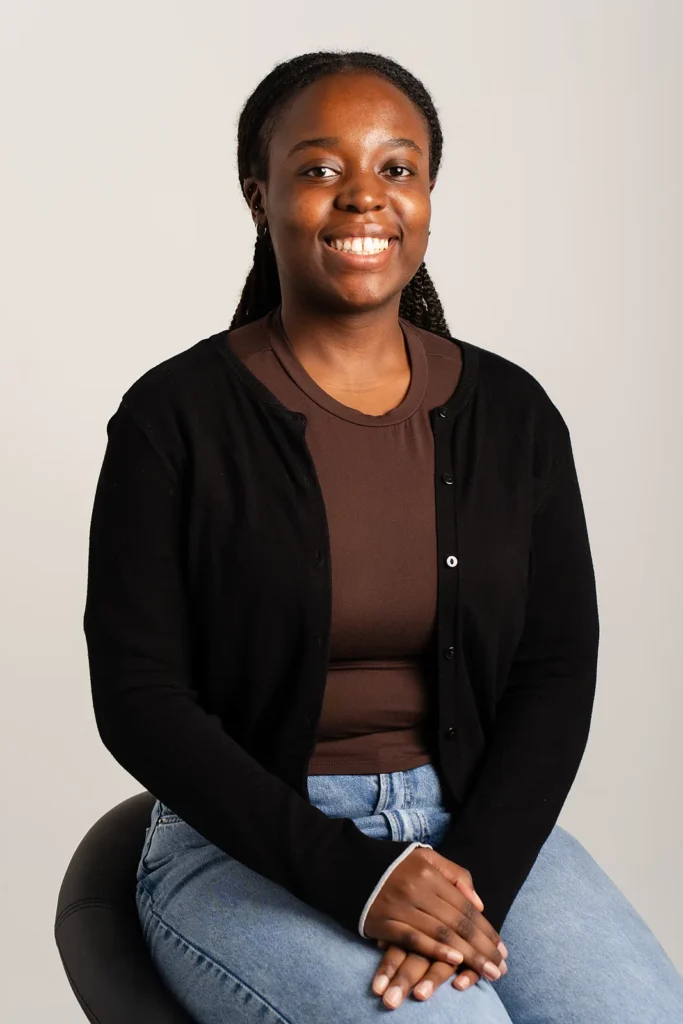Professional corporate headshot of a smiling woman with braided hair, wearing a black cardigan over a brown top, against a neutral studio background.