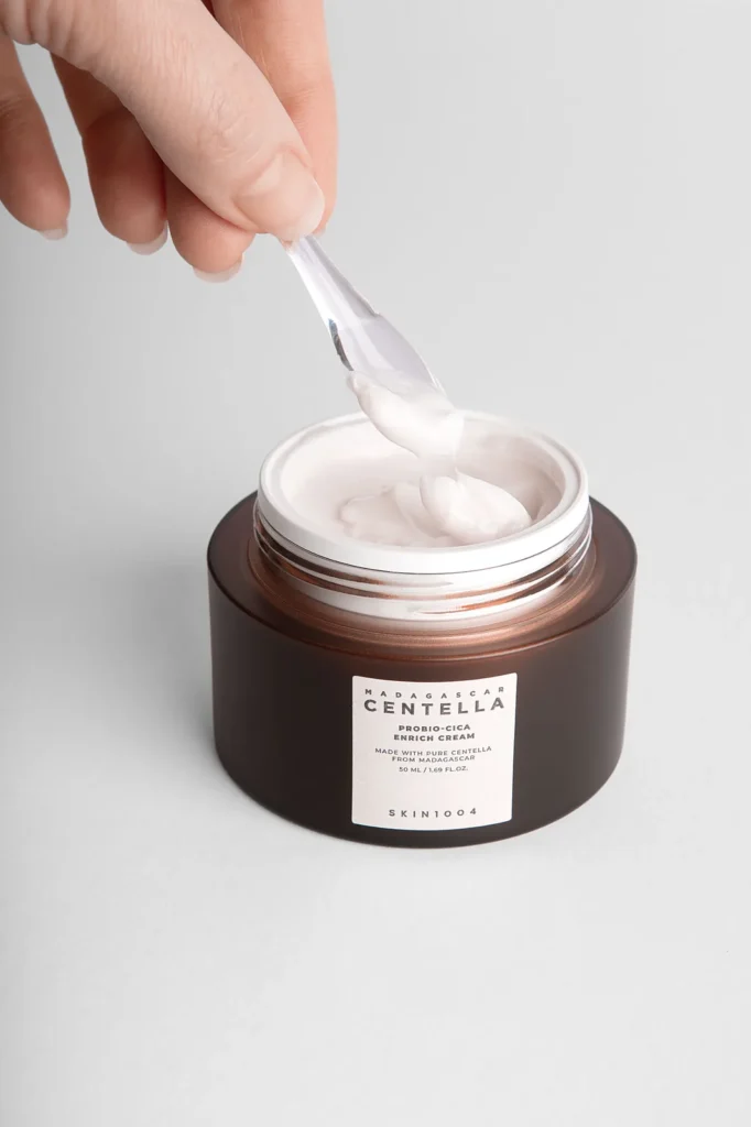 Hand lifting face cream from an amber jar with a clear spatula, lifestyle product photography for a skincare brand.
